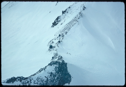 An aerial photo of a string of small buildings on a rocky ridge with steep snowy slopes on either side.