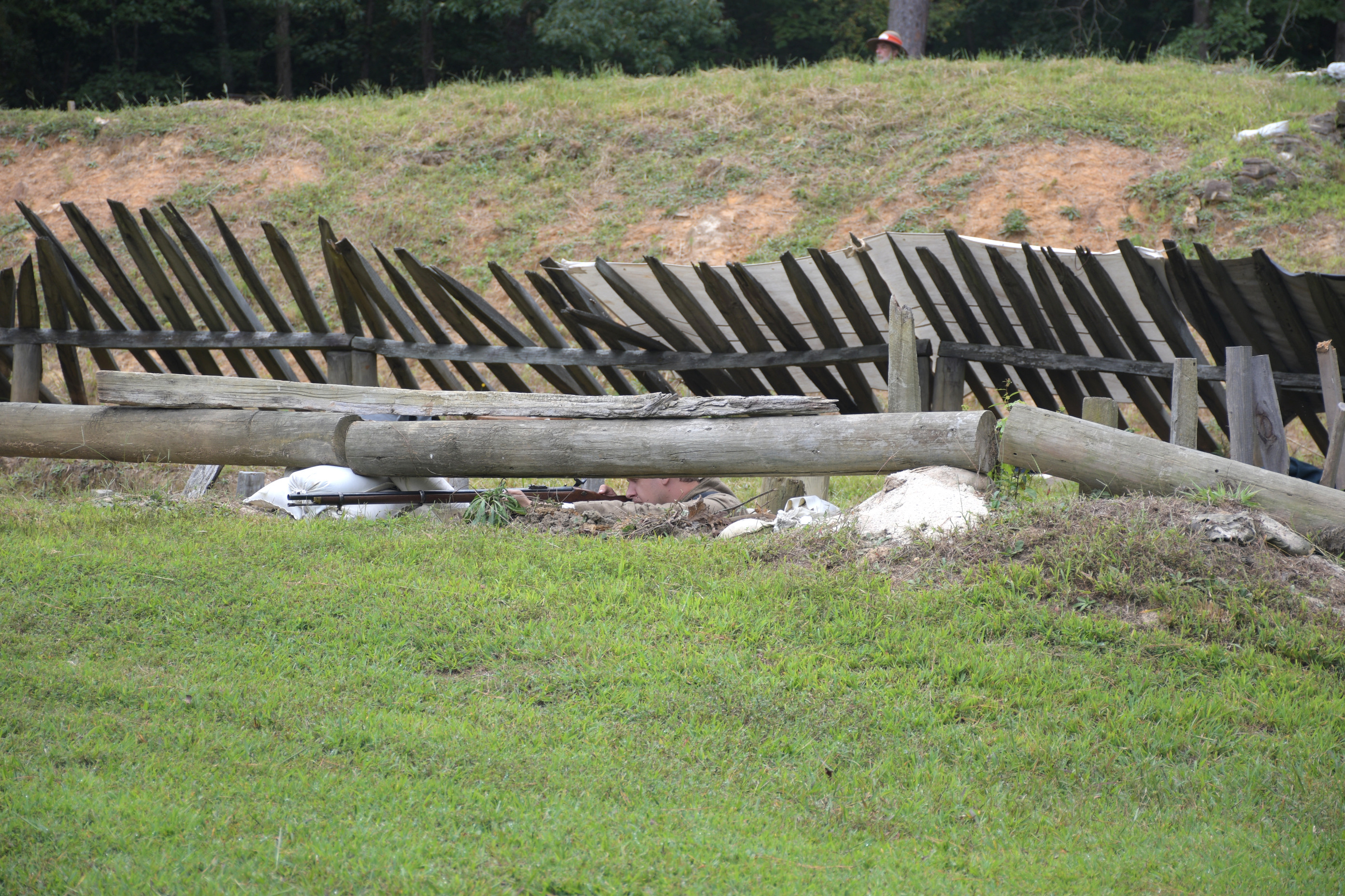 A white man wearing Confederate uniform aiming a rifle just visible behind a head-log.