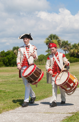 Two living historians, a young man and a younger boy, walk side-by-side on a paved path while playing large, traditional military drums. They wear matching white and red 18th-century uniforms with distinctive hats. In the background is a blue sky, green trees and bushes and green grass. 