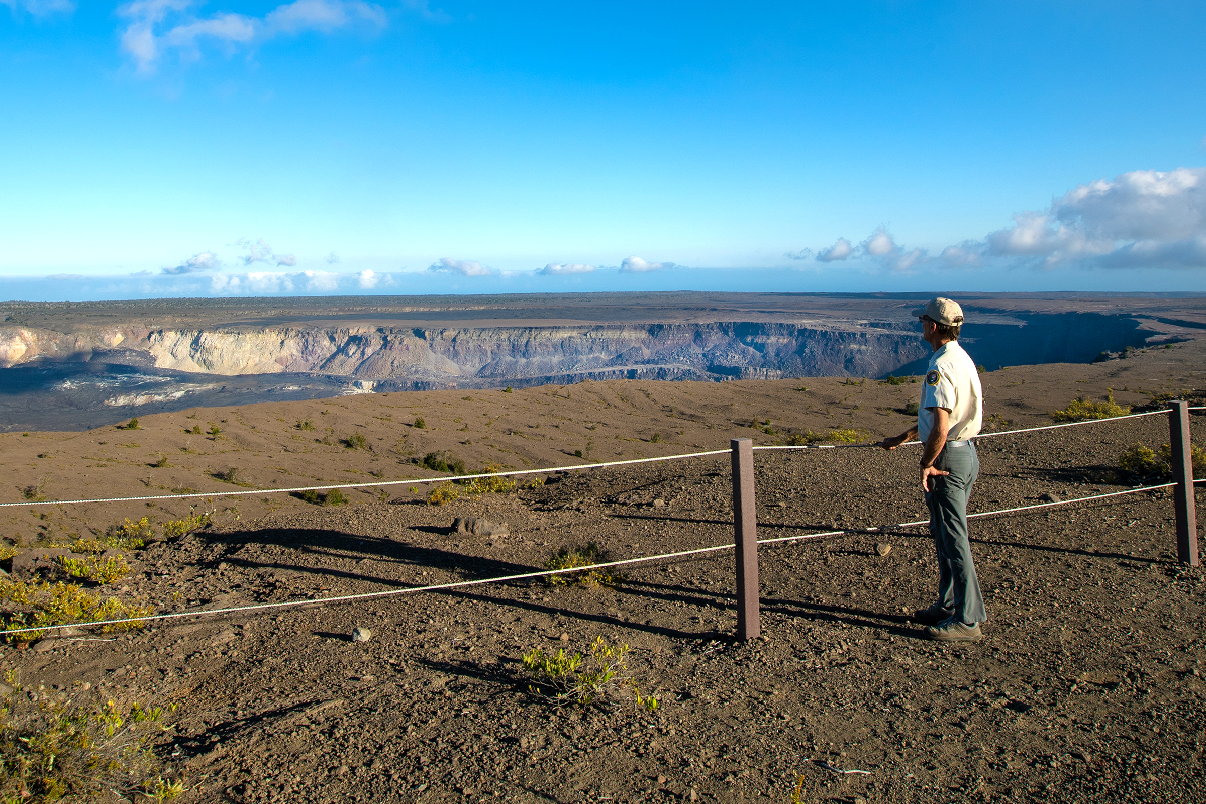 A man in a cap with his hand on a railing looks out across a vast volcanic summit
