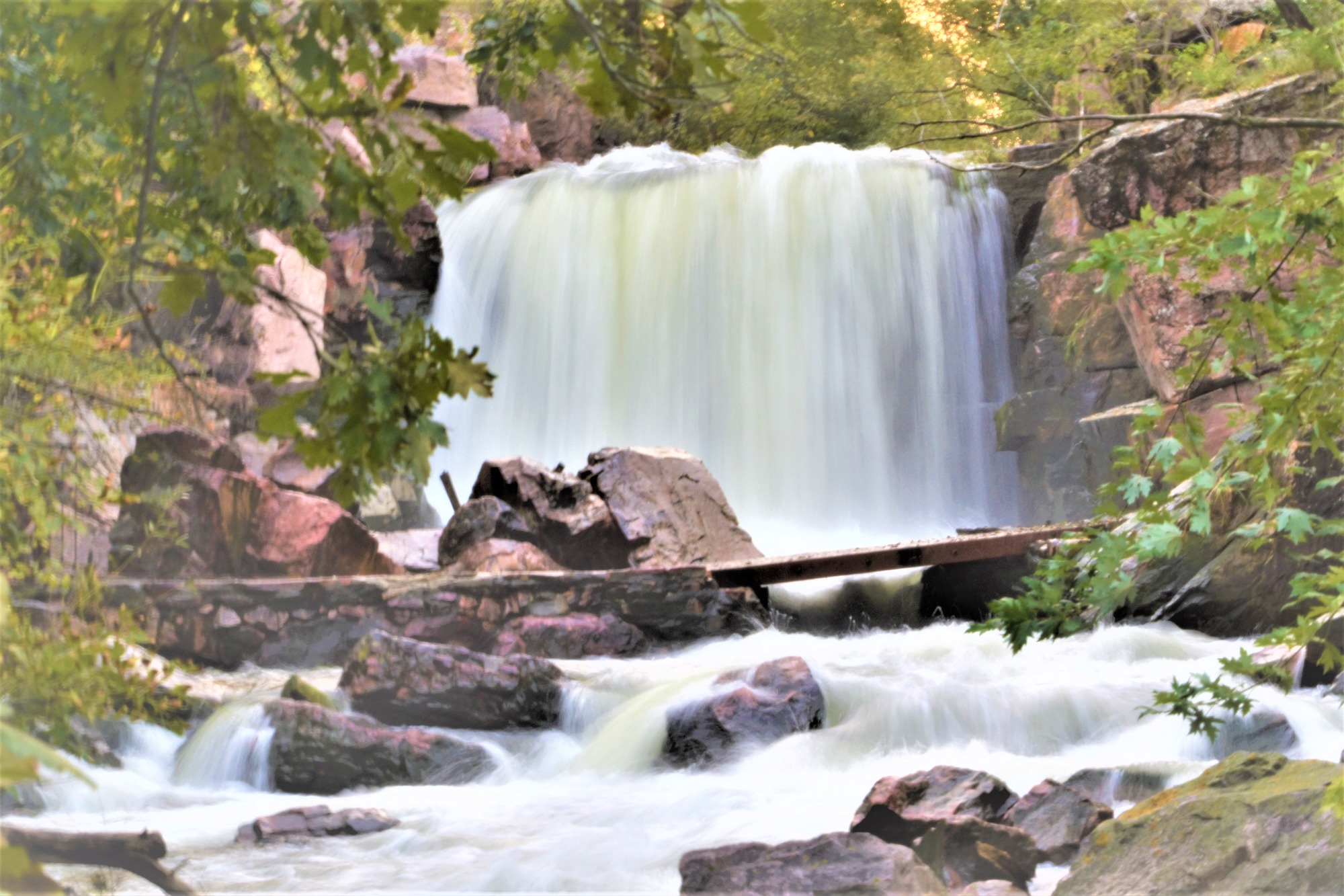 Waterfall behind a creek which is crossed by a footbridge. Trees frame the image. 