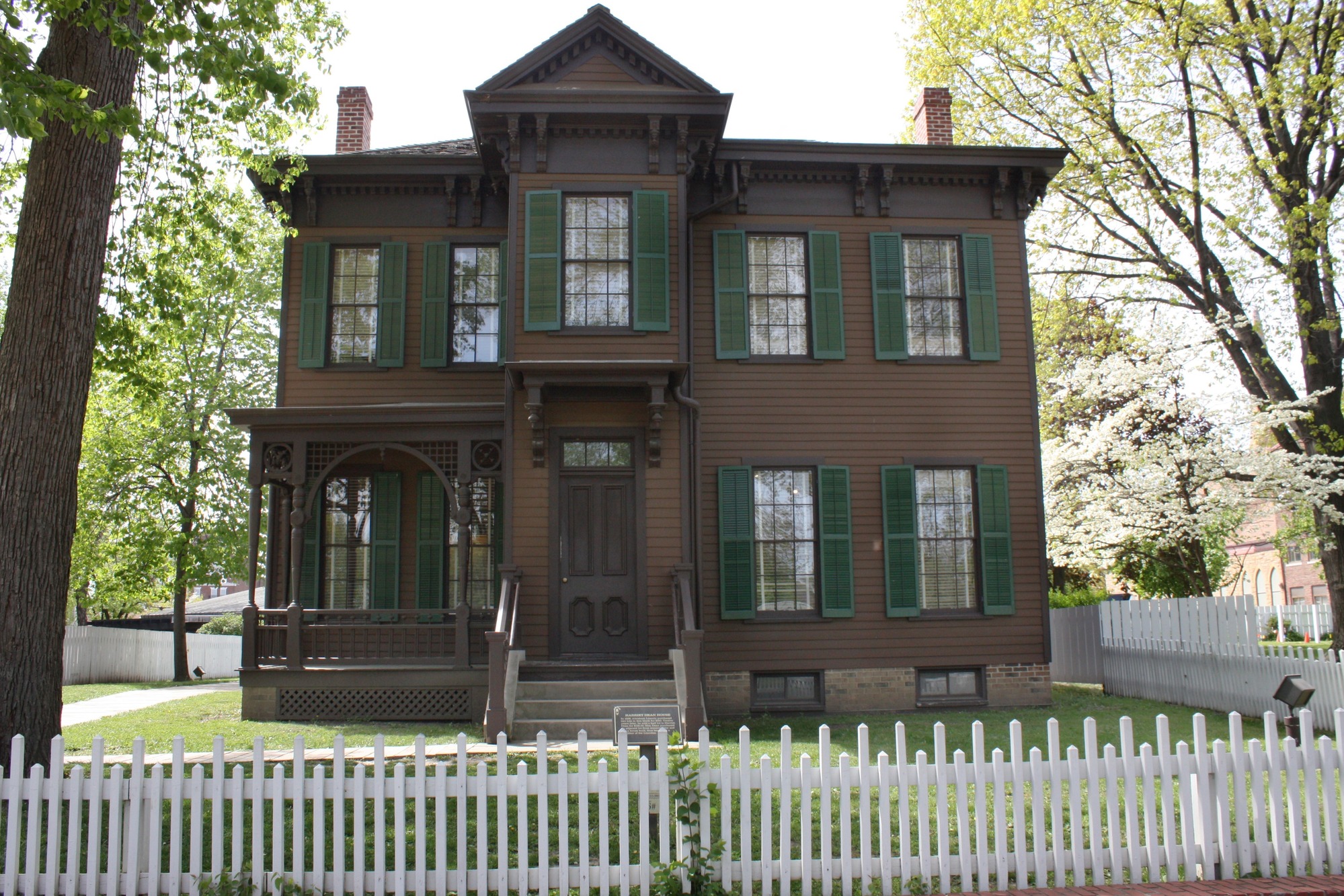 A two-story wooden house, painted dark brown with green shutters, surrounded by a white picket fence.