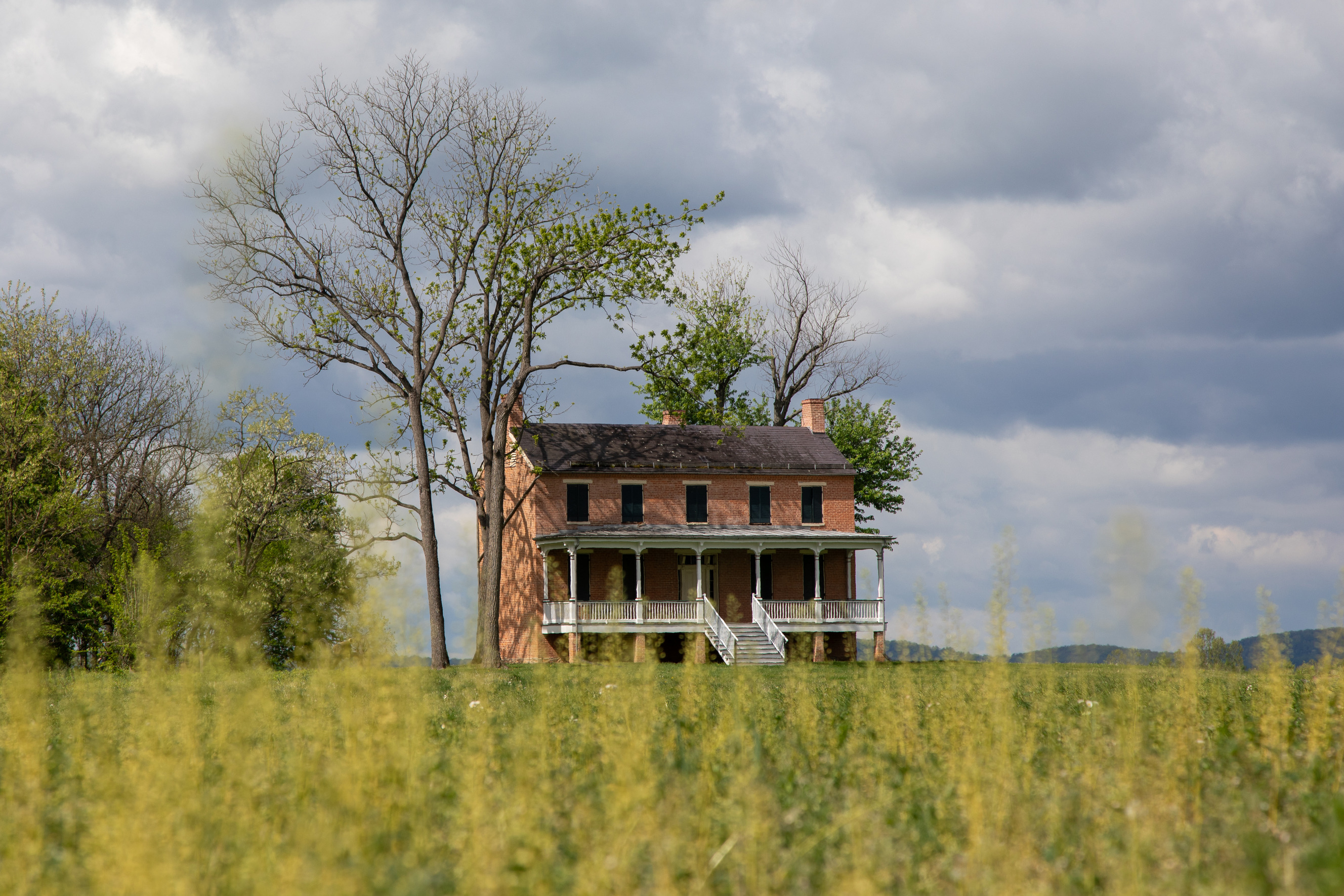 A grassy field and a two-story brick house with a porch in the distance.