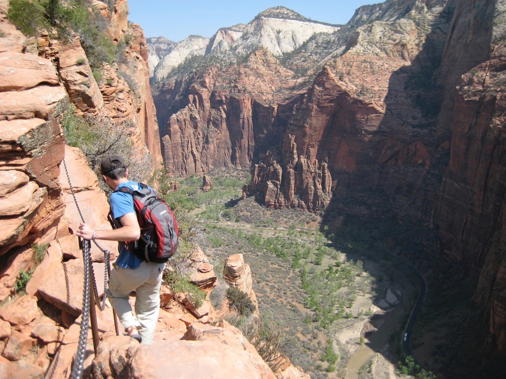A hiker holds onto a chain railing as they walk a narrow ledge with a large canyon below. 