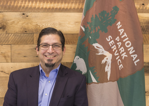 Man stands in front of a wooden wall and NPS flag.