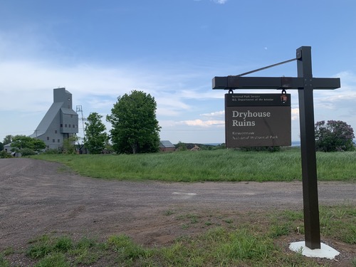 At the corner of a gravel road is a NPS sign that ready Dry House Ruins.  Across the street in the background is the Quincy shaft house. 