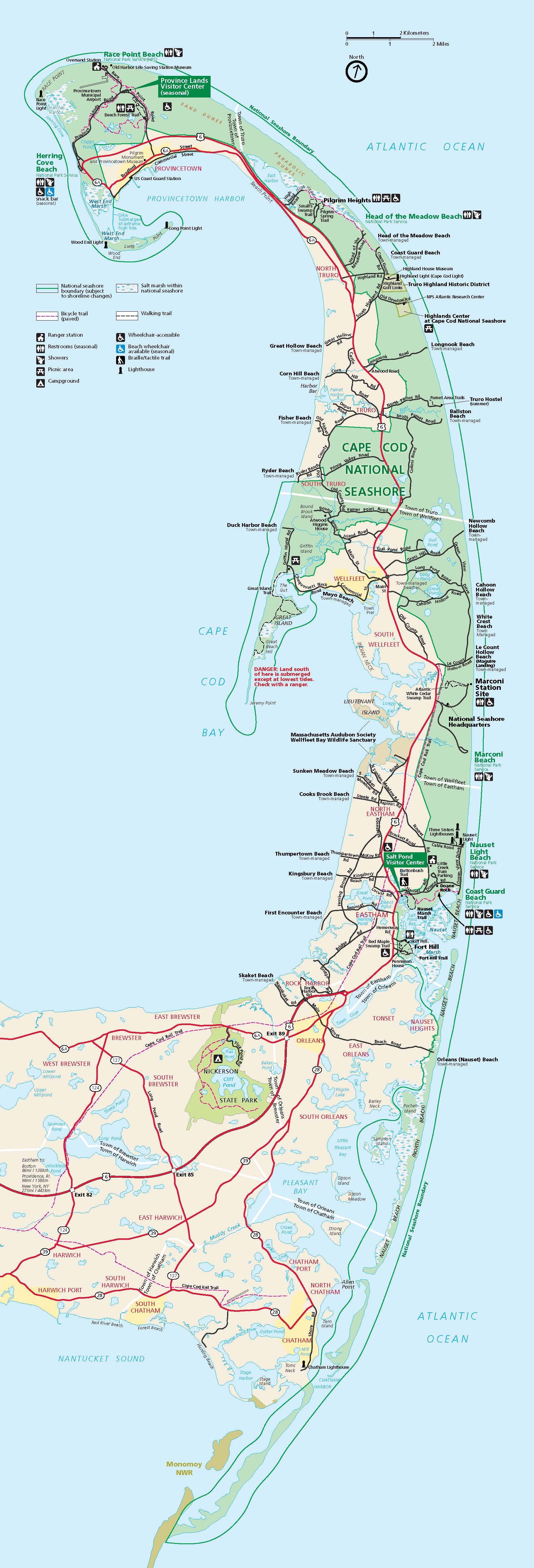 Map of Cape Cod National Seashore in Massachusetts. A backward L-shaped peninsula runs vertically up this tall map. The bottom, horizontal section, which reaches in from the left edge, is beige and the Cape Cod National Seashore is shown in green. The water is blue and a network of roads is shown in black and red.