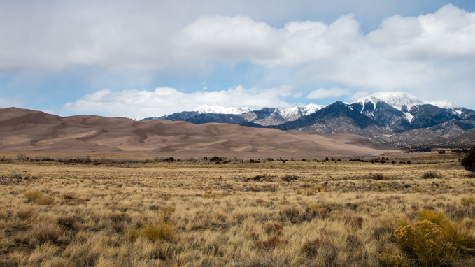 A large field of shrubs and grasses leads toward the sand dunes. Behind the sand dunes the snow-capped Sangre de Cristo Mountains rise above the valley floor. 
