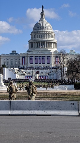 U.S. military standing watching the U.S. Capitol building, which has a large crowd gathered on its various levels and is decorated with large U.S. flags and red, white, and blue banners.