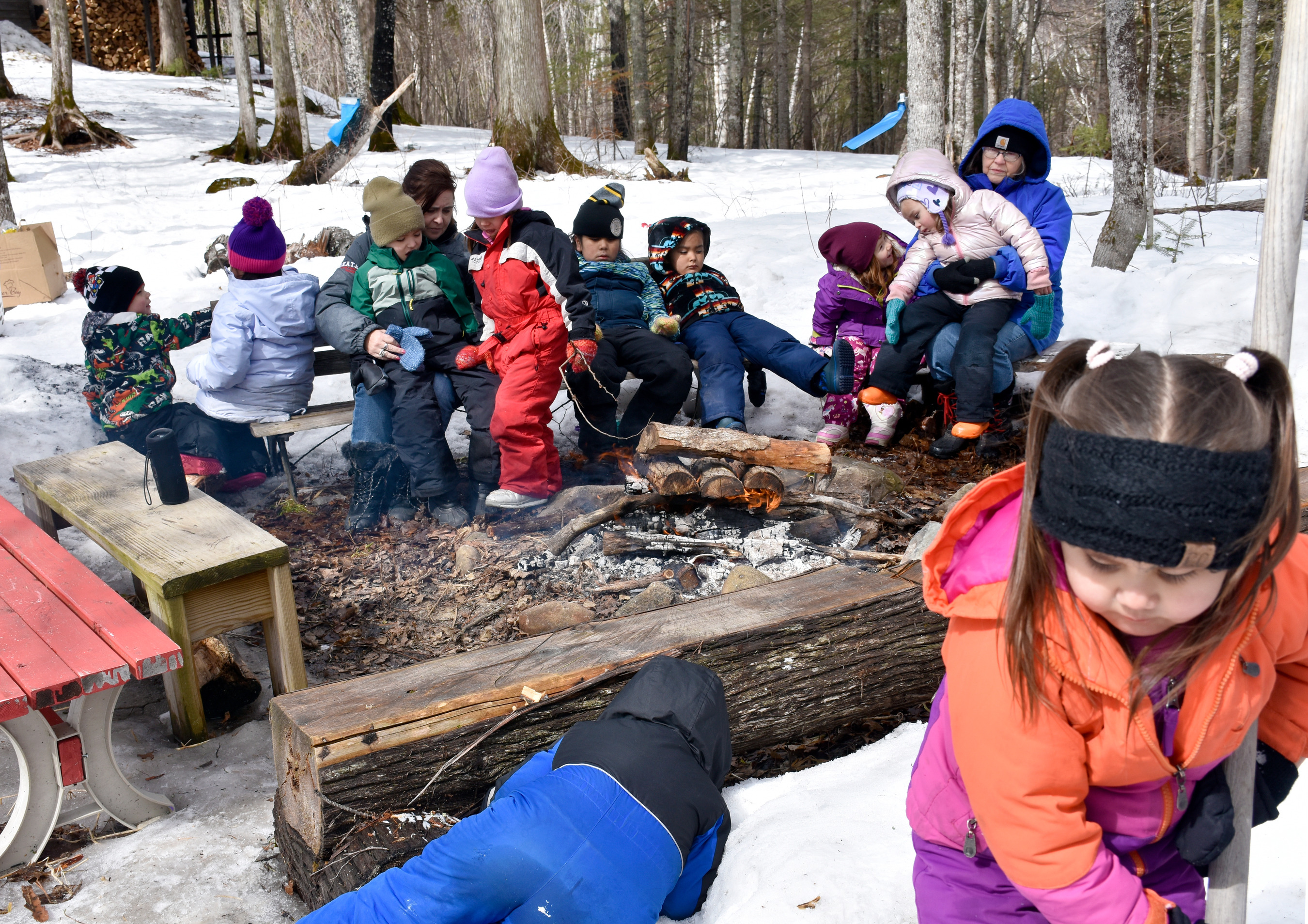 Ten children and two adults in winter clothes sit on benches near a fire. One child reaches for something under a log and one shovels snow.