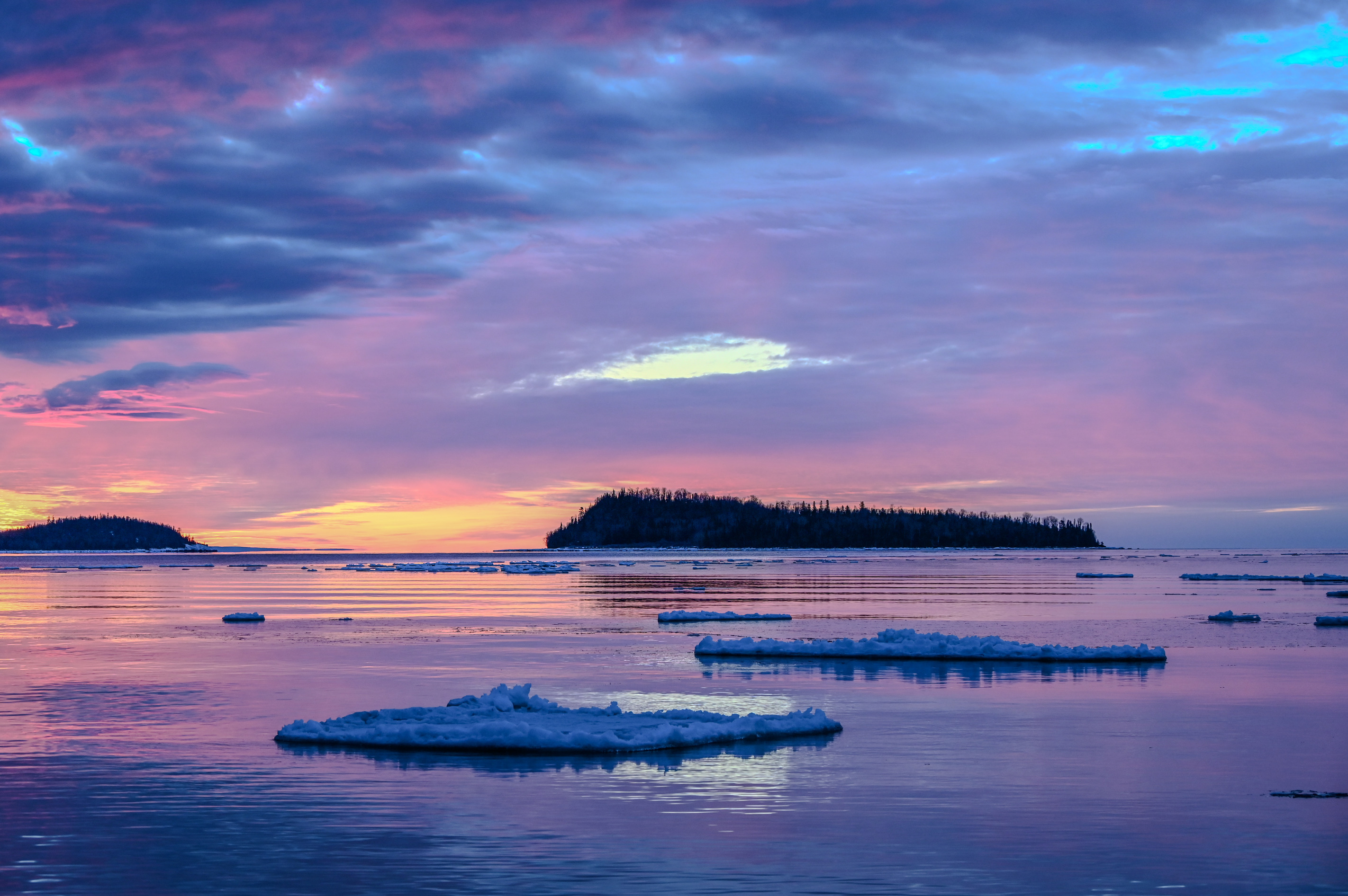 Ice floats in the water of Grand Portage bay with Grand Portage Island in the distance. A colorful purple, pink, and orange sunrise reflects in the calm waters.