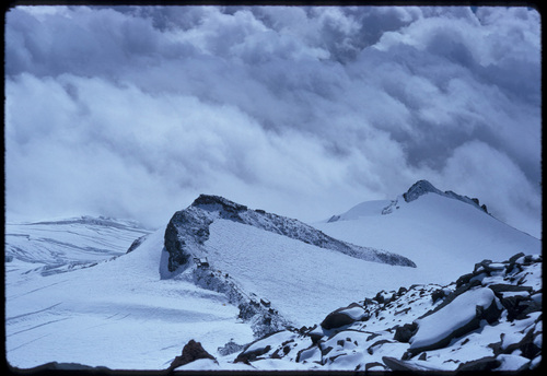 Looking down a steep snowy slope, a string of tiny buildings perch along a rocky ridge that juts out in a curve from the slope, with clouds filling the valley below the ridge.