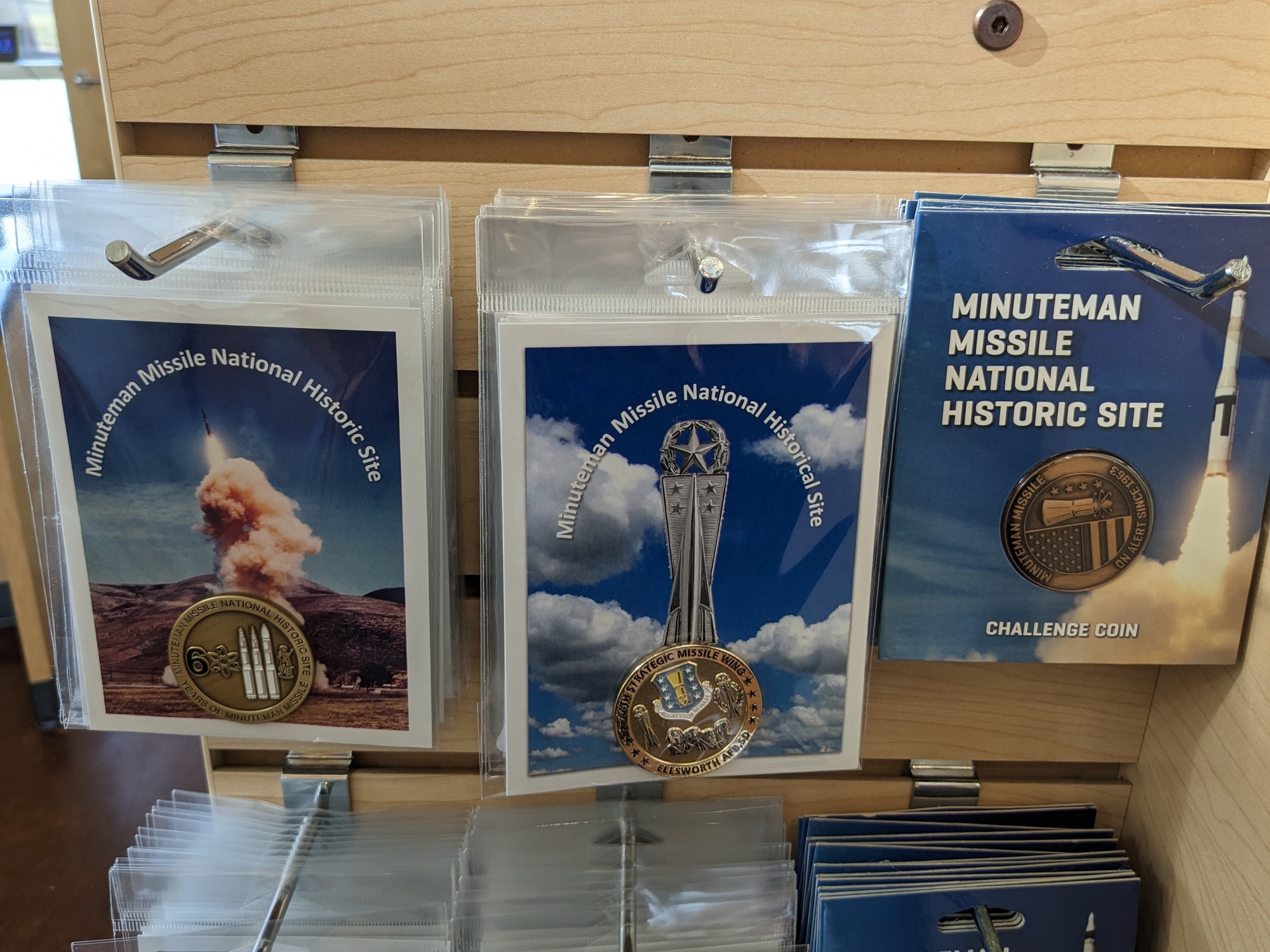 Challenge coins hanging on a rack. One is a dull colored brass with image of three missiles and year "60". One is a shinier gold color with the 44th Strategic Missile Wing emblem, and one is a copper/bronze color with the park logo.  