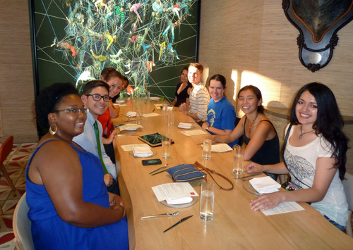 a group of 10 young adults sit at a long wooden table at a restaurant and smile for the camera at the end of the table.