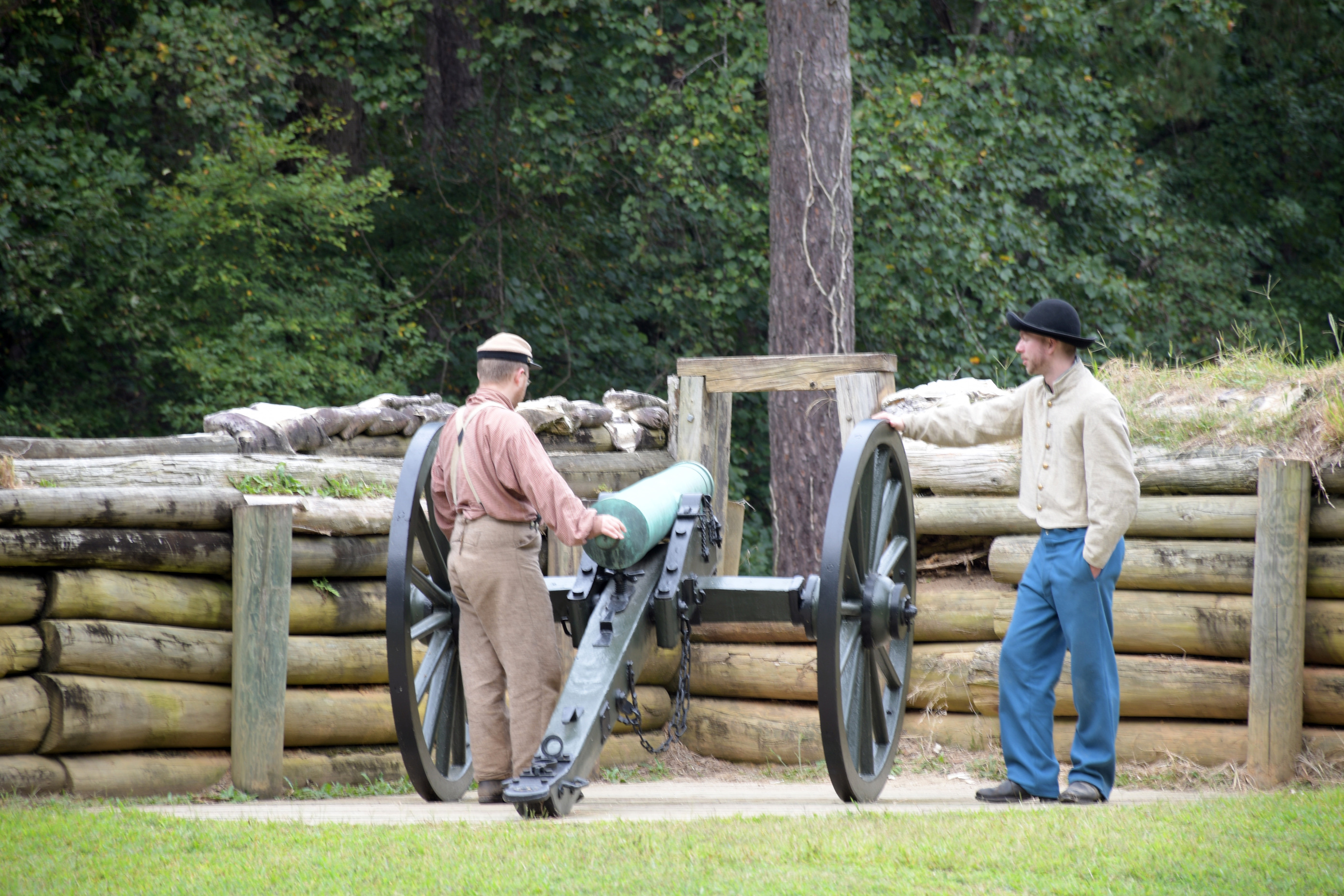 Two white men in Confederate uniforms stand looking at a cannon with wooden retaining wall supporting earthworks in the background.