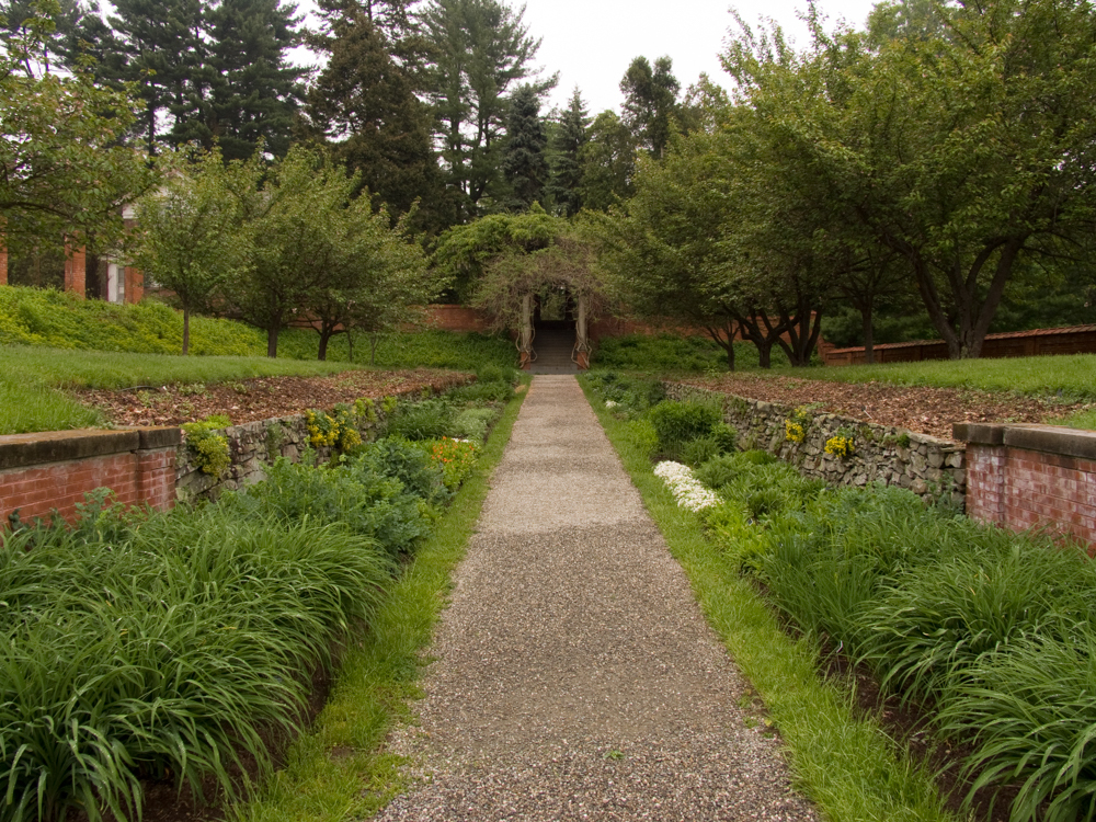 A gravel garden path flanked by brick and stone walls and flowering plants.