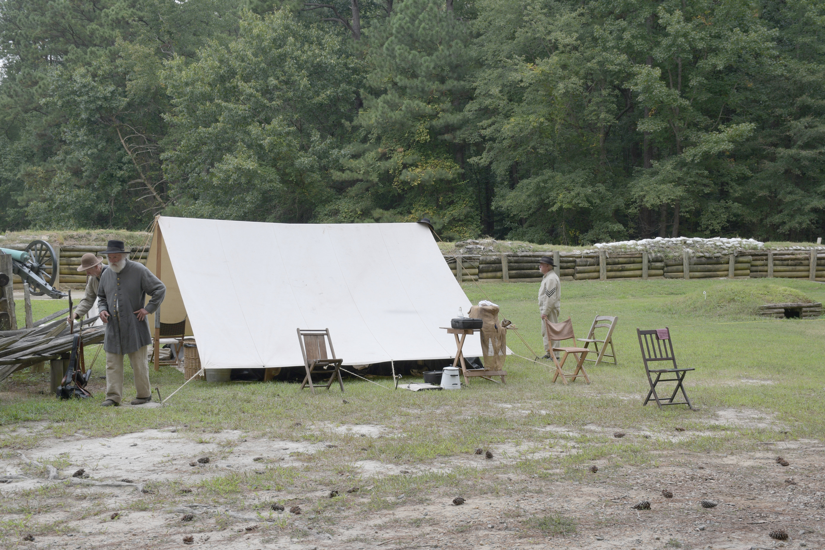 Three white men wearing Confederate uniforms walkaround a white canvas tent several chairs and a table outside the tent. A wooden retaining wall supports earthworks in the background.