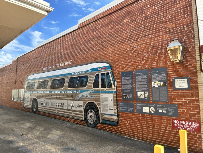 A mural of a Greyhound bus is painted on the side of a building. Surrounding it and covering it are educational panels about the Freedom Riders. Above the mural are the words "Could You Get On the Bus?". An eternal flame in a gas streetlight hangs above and to the right of the mural and educational panels. 