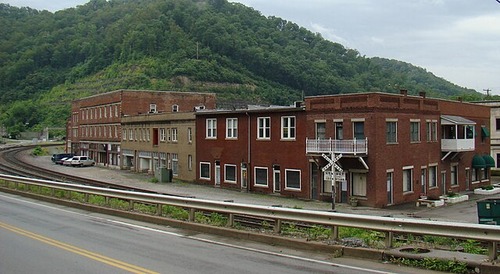 The foreground shows part of a modern two-way road. Railroad tracks run parallel to the road. The railroad tracks sit between the road and a row of two to three story brick buildings. Altogether, the landscape shows a small, nondescript mountain town.