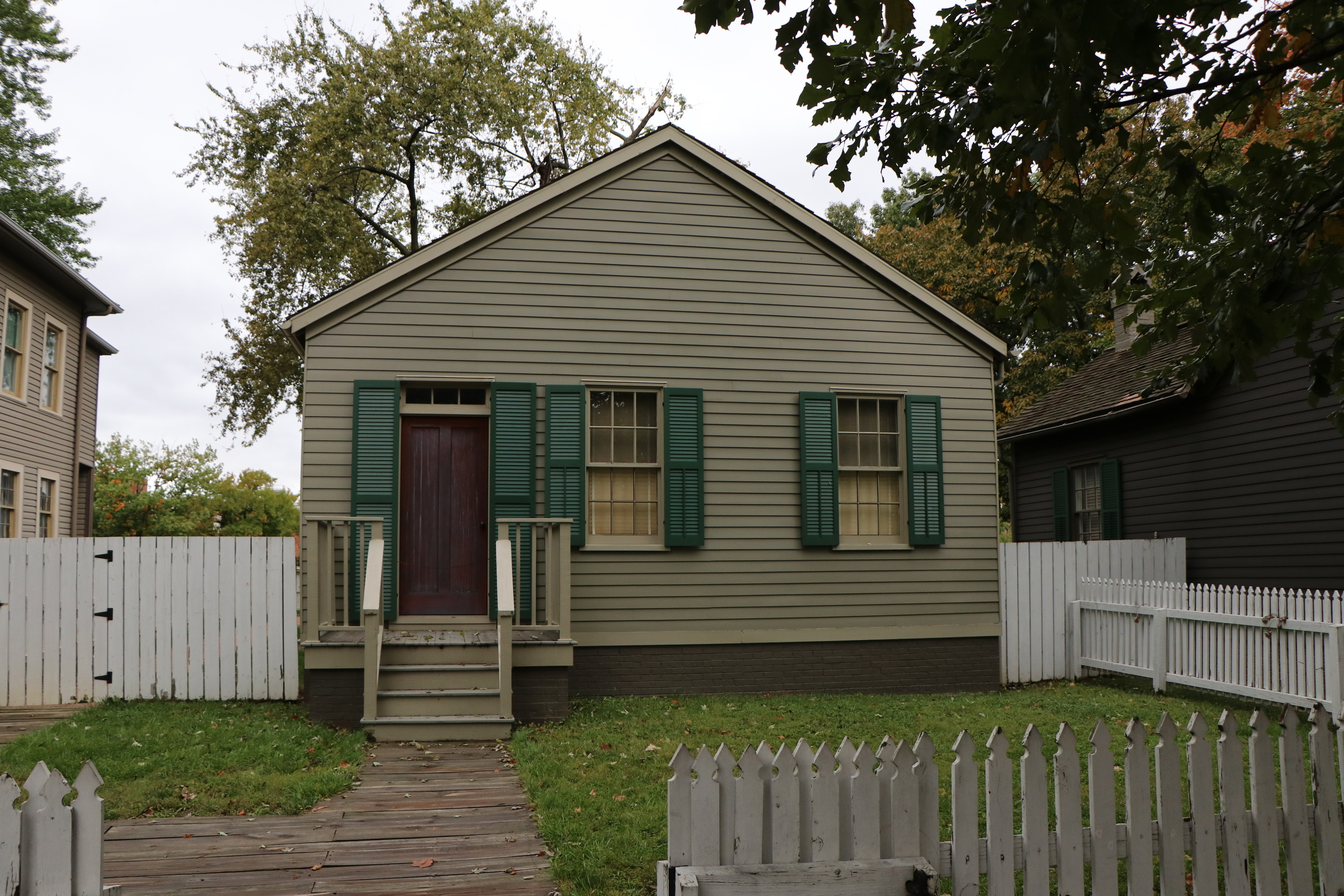 A one-story green house with green shutters and a fenced-in front yard.