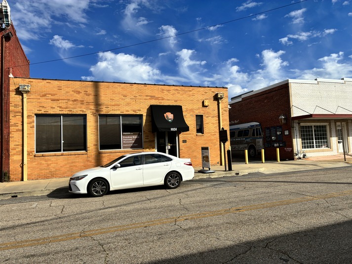 A tall, one-story brick building with large windows and a double door out front. To the right is an alleyway with a mural of a Greyhound bus painted on the wall. Further right is another large brick building. 