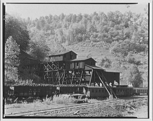 A mountain-side landscape with tree-lined hills. The center of the image is a valley where a large frame structure stands on stilts and trusses. The structure has two enclosed, frame buildings with gabled rooves on top of the stilts. They are built at different heights but are in line with one another by their side elevations. The entire structure gradually decreases in height in a line towards the right of the photo. Where it terminates in additional frame, compartment-like components. Right next to the structure are railroad tracks. 