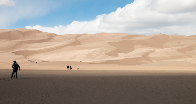 Visitors walk across the sand flats that lead to the dune field. The sun is shining down on the dunes. The sand flat is in the shade. 