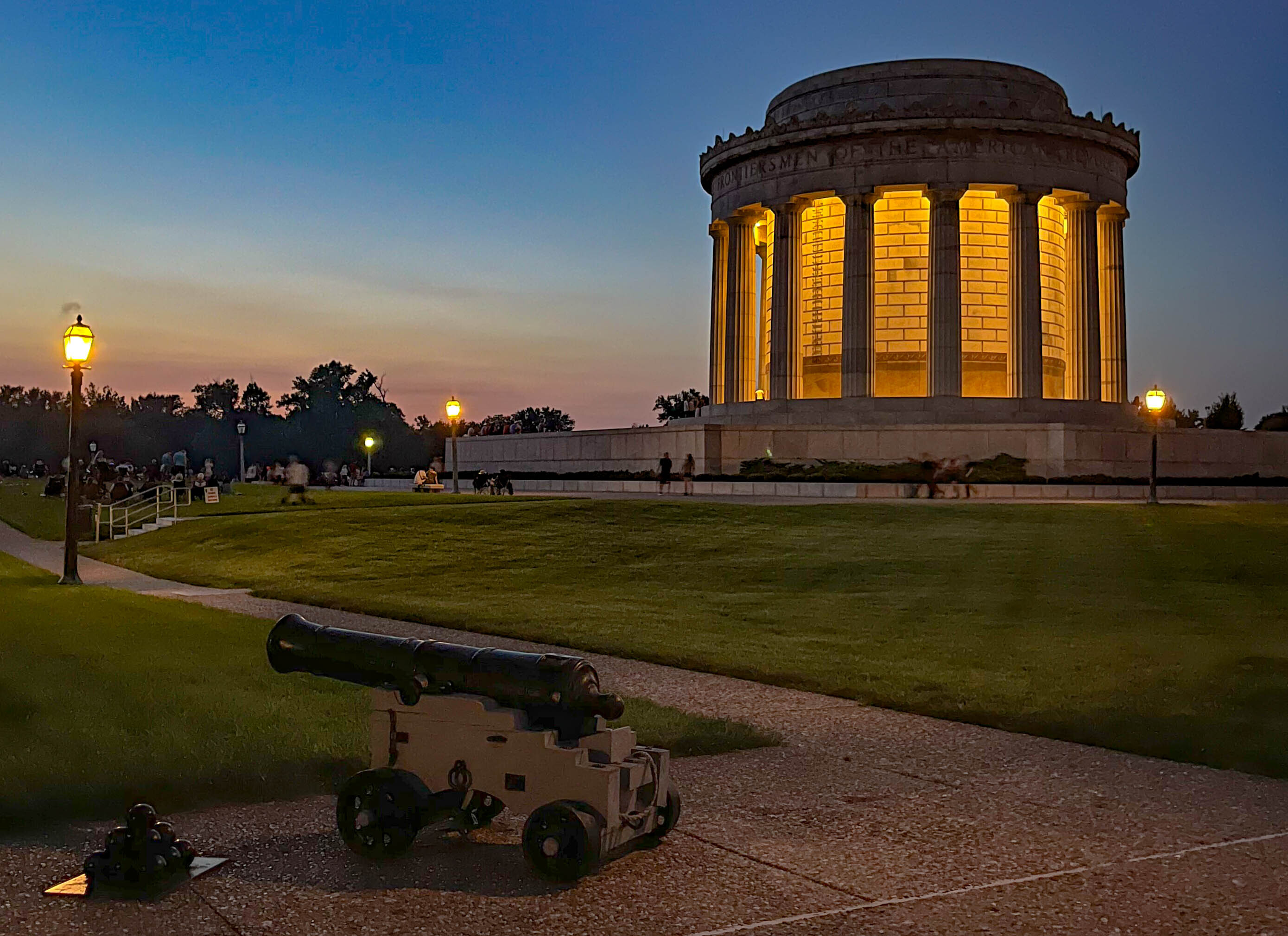 Large circular classical-style memorial building with prominent columns at night with a cannon and cannonballs in the forefront