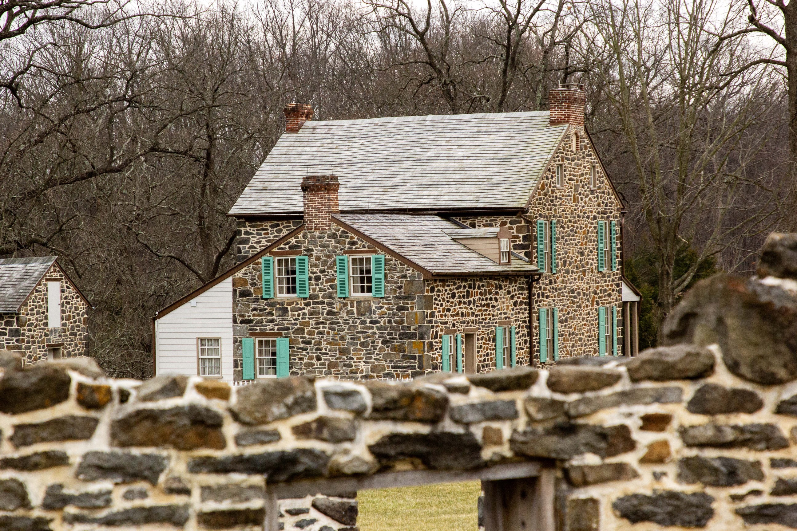 A two-story stone farmhouse with green shutters. Behind the farmhouse is a wall of bare trees. In the foreground are the remains of a stone wall. 