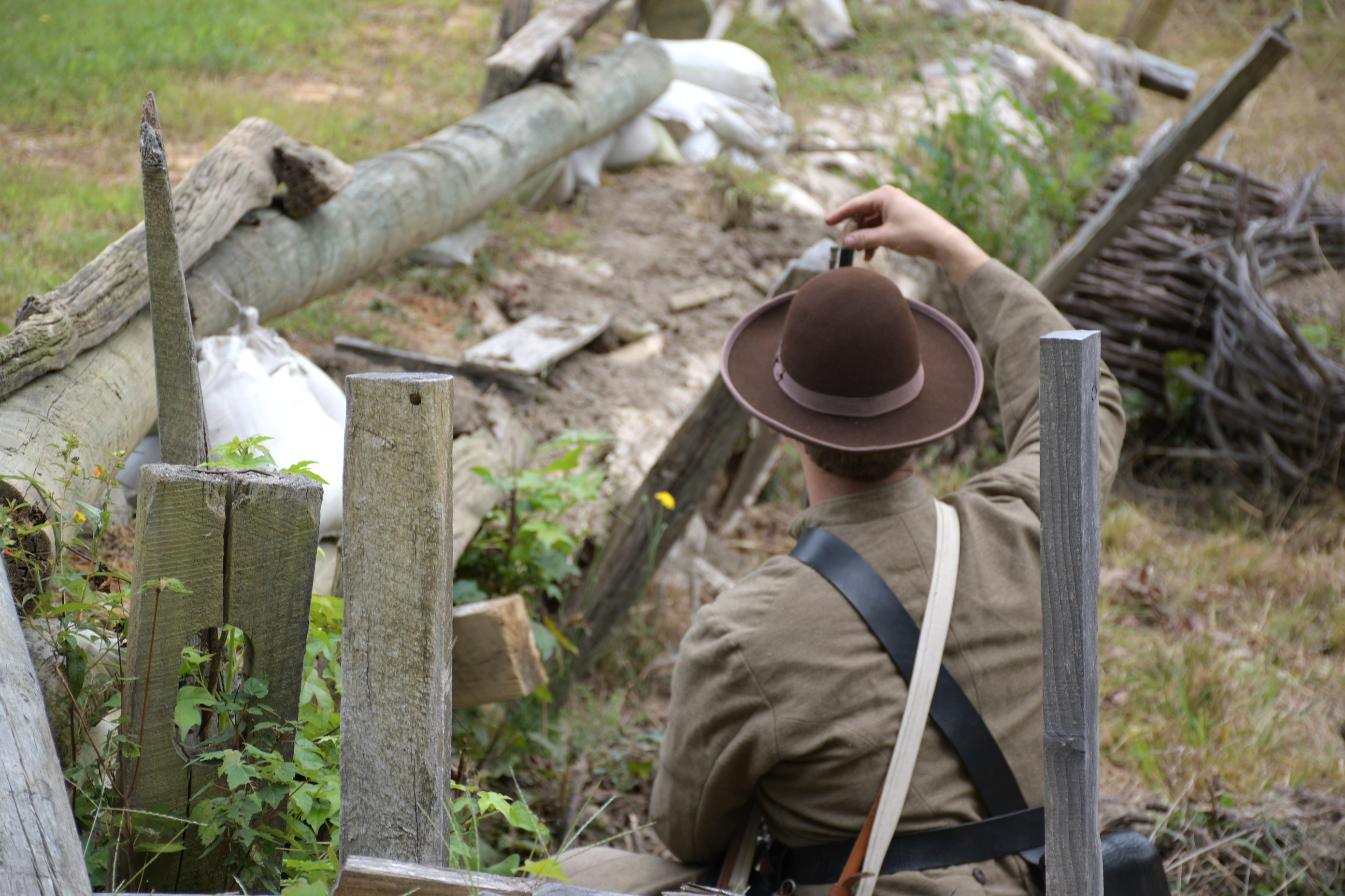 The back of a white man wearing a Confederate uniform and brown hat kneels behind earthworks - loads a rifle.