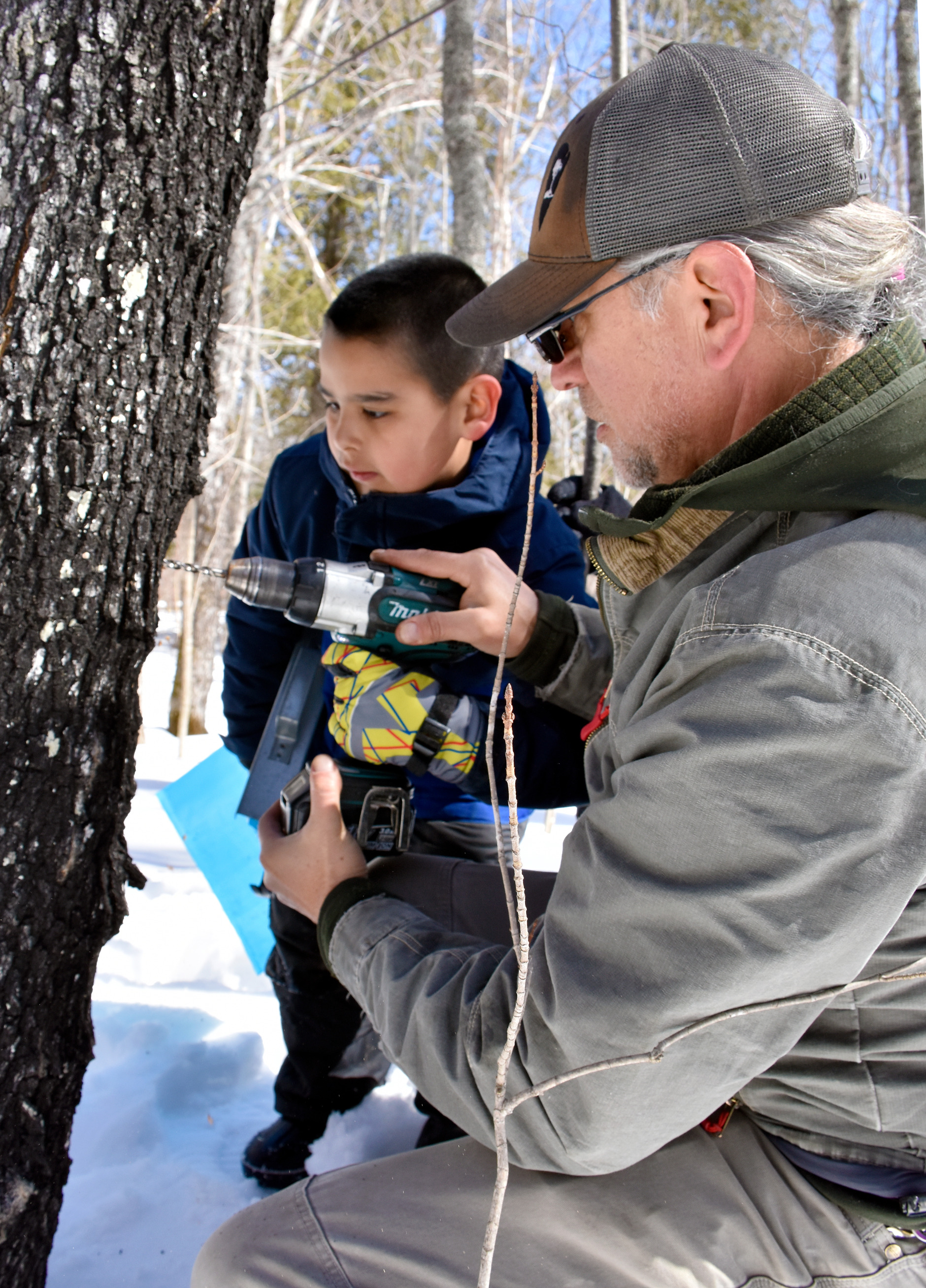 An adult wearing grey clothing holds a drill so that a child can press the trigger to make a hole.