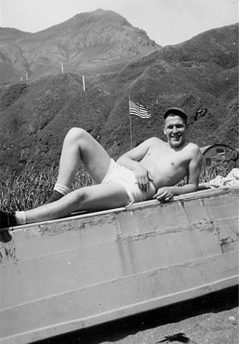 A shirtless man lays down in front of mountains and an American Flag. The photo is black and white.