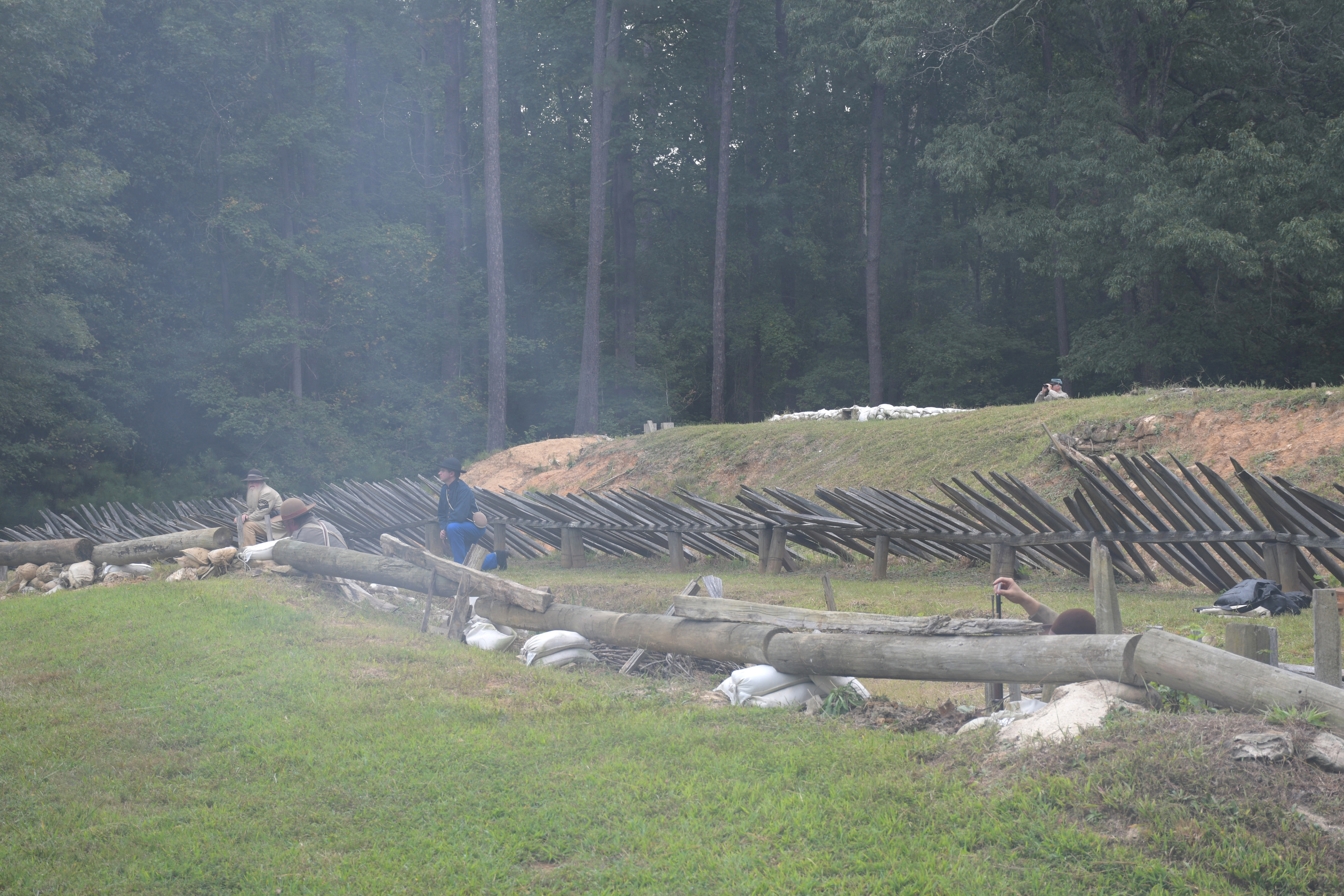 The smoke of creates a haze in the air.  Five white men wearing Confederate uniforms four just visible behind head-logs in the foreground and one visible above earthwork in the background.
