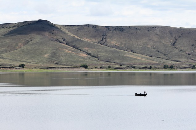 A small fishing boat with a person standing on it floats in a large body of water. Mesas are in the background.
