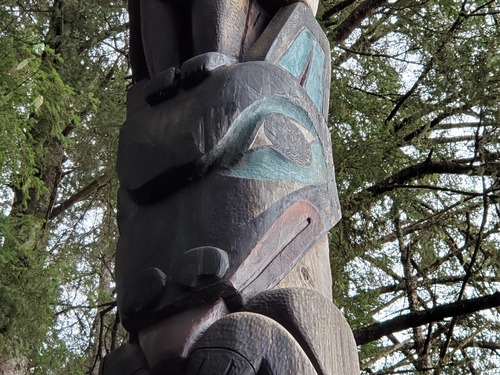 Close up detail of a carved wolf face on a totem pole. the face is black with red tongue and lips  and blue green around the eyes and inside the ear. 