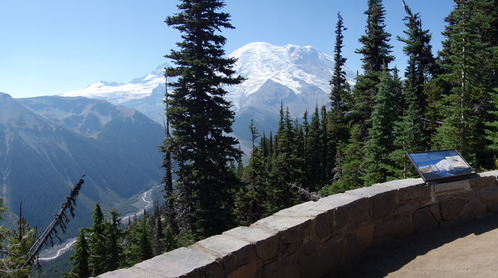 An exhibit panel attached to a low rock wall overlooks a view of a glaciated mountain and a wide forested river valley. 