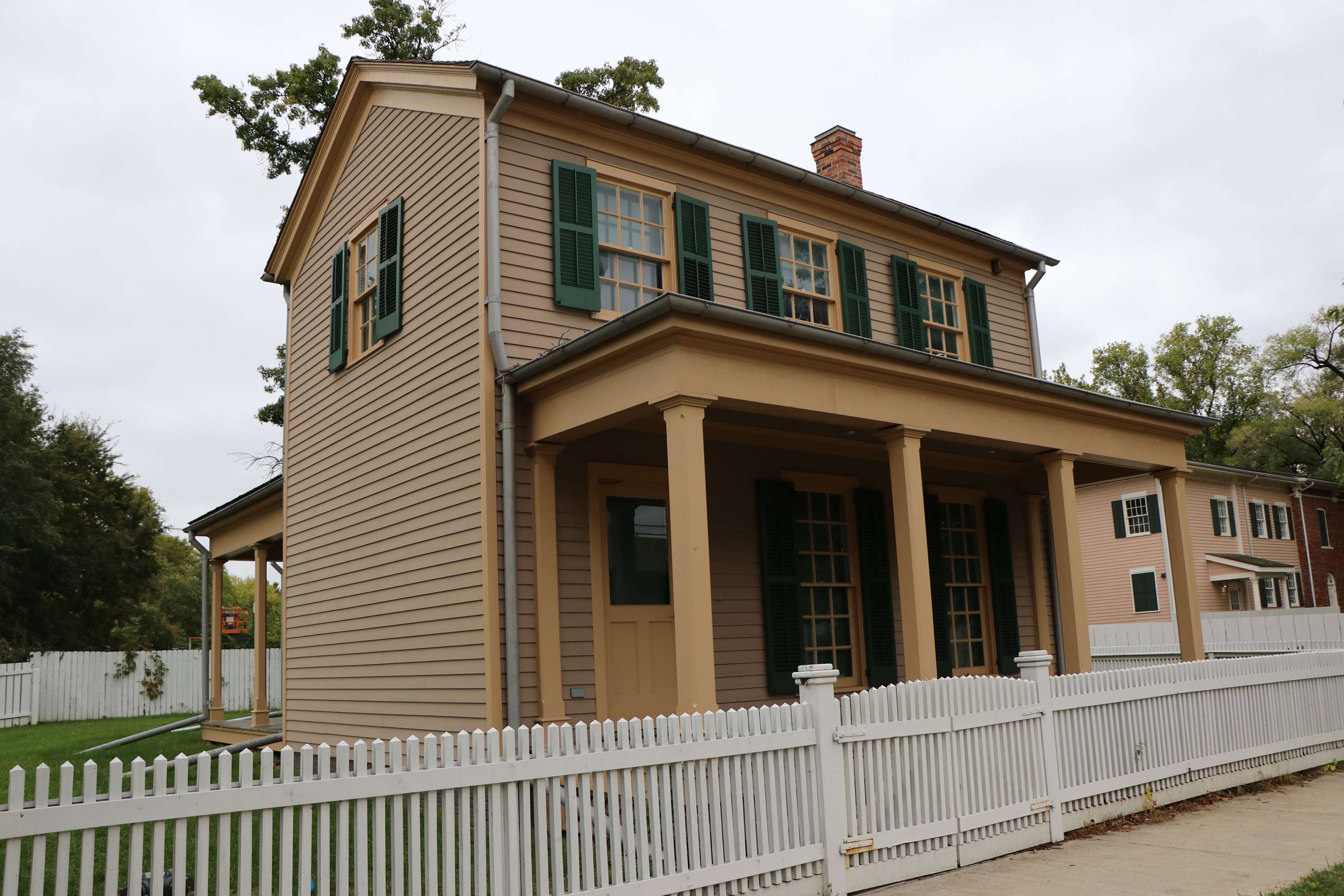 A two-story tan house with green shutters and a white fence in front of it.
