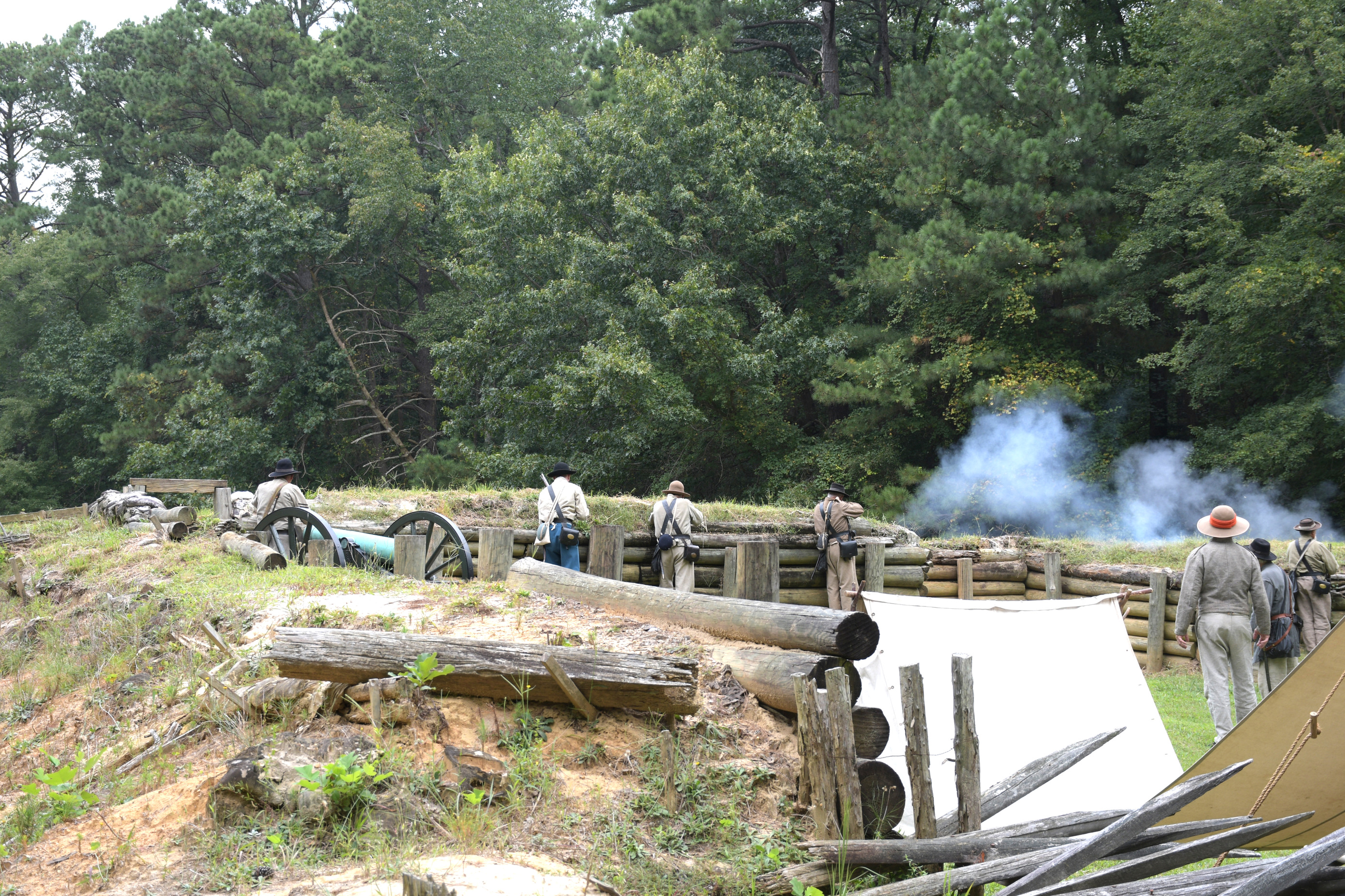 Seven white men wearing Confederate uniforms firing rifles from defensive positions behind earthworks.