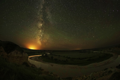 The Milky Way shines bright in the night sky above the Little Missouri River in Wind Canyon, Theodore Roosevelt National Park, North Dakota. Light pollution / sky glow is visible at the horizon and base of the Milky Way.