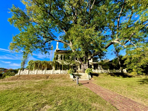 Historic two-story white home with a prominent covered wrapped porch and a brick walkway, interpretive signs, stairs, and tall tree in front