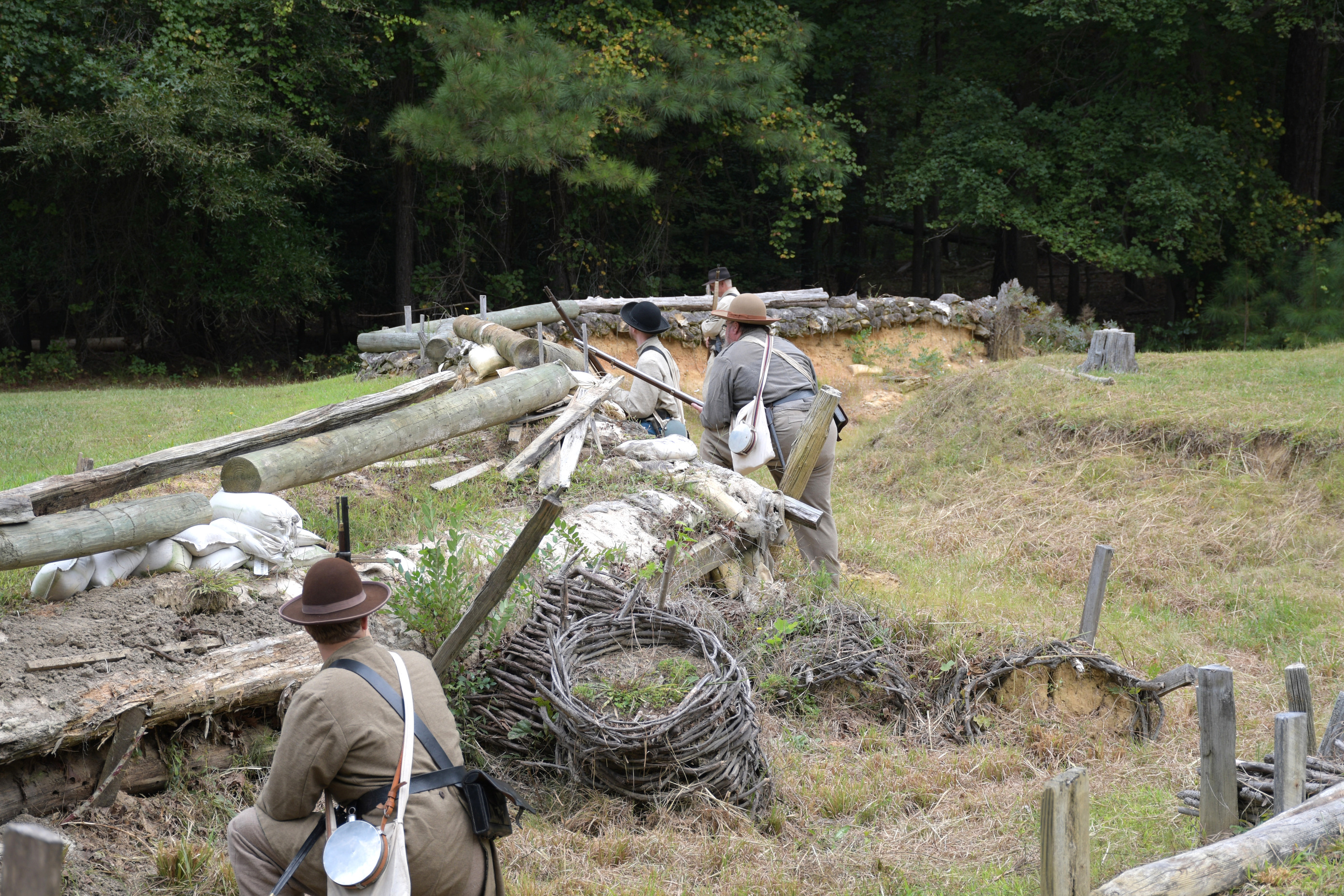 Four white men wearing Confederate uniforms carrying rifles take position behind earthworks.