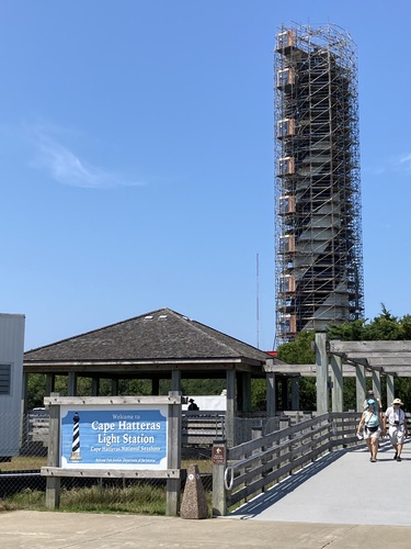 Cape Hatteras Light Station sign in foreground and lighthouse with scaffolding sits in the background on a blue sky day.