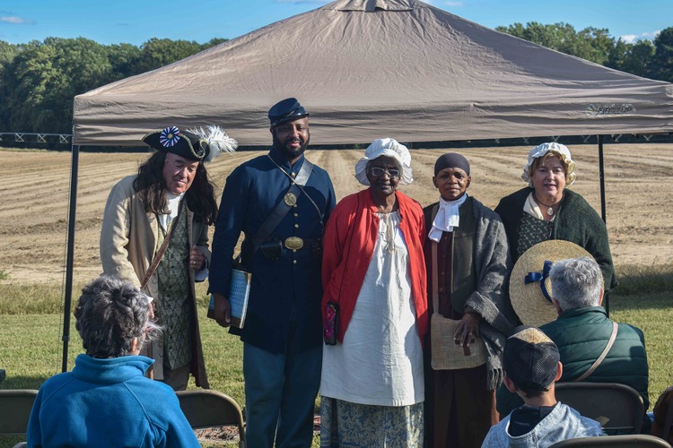 Five adults wearing 18th and 19th century clothing can be seen in the center of the image standing side-by-side. A tan tent, open field, and tree line can be seen in the background. Seated on lookers can be seen in the foreground.