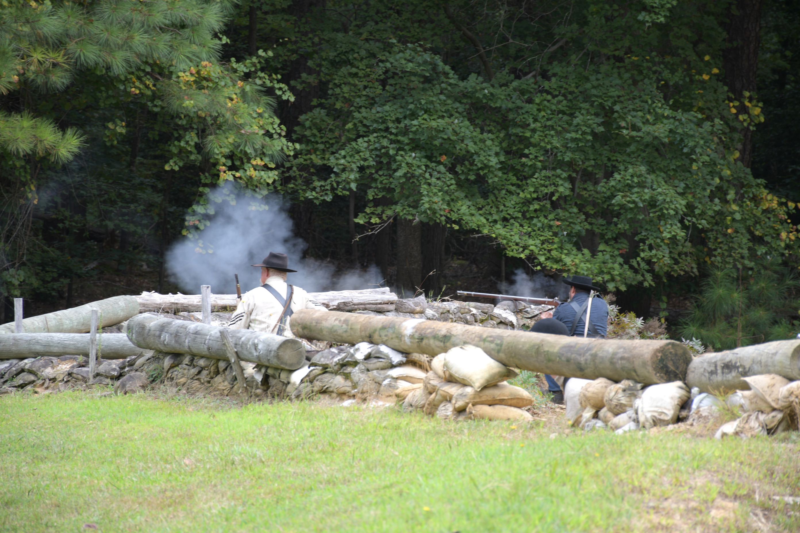 Two white men wearing Confederate uniforms firing rifles from defensive positions behind earthworks.
