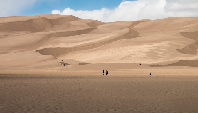 Park visitors walk across the sandy flat and up the smaller dunes at the beginning of the dune field. The sun is shining on most of the dunes while other parts of the dunes are in shade. 