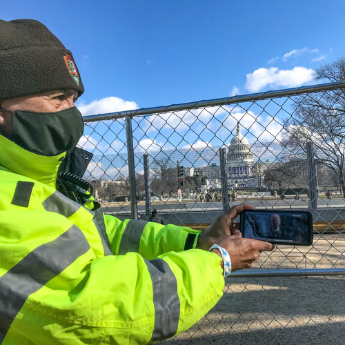 A National Park Service employee holding up a cell phone displaying President Biden giving his inaugural address with the U.S. Capitol building in the background behind fence lines