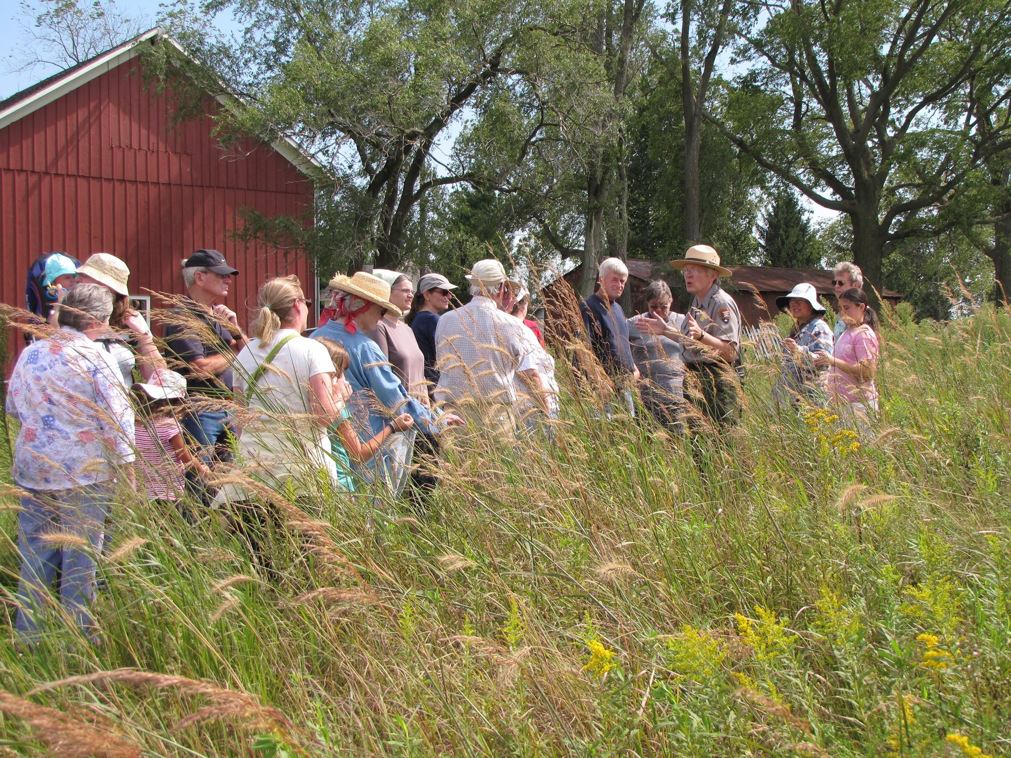 A park ranger speaks to a group of people by a red barn near a prairie trail.