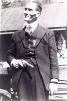 A three-quarter frame black and white portrait of a man looking to the left of frame. He is wearing a full suit and holds a small handgun pointing down.  
