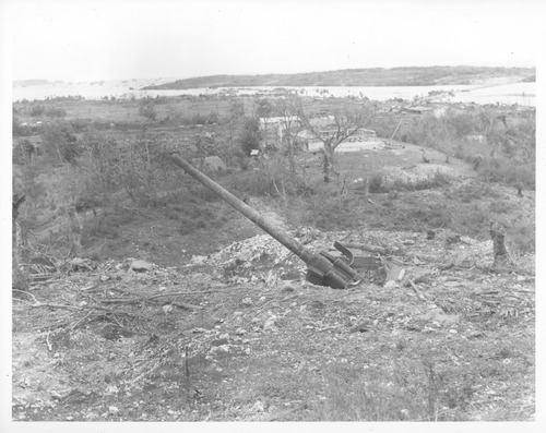 Large gun pointing towards the sky. The ground around the gun has been damaged from bombings and in the background there are the ruins of buildings. 
