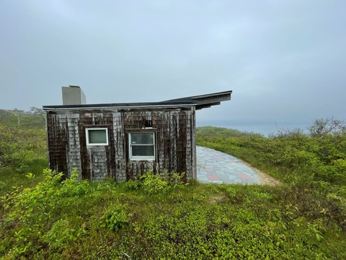 Exterior view of home looking out onto the Atlantic Ocean. Shingled one story home with curved patio surrounded by greenery.